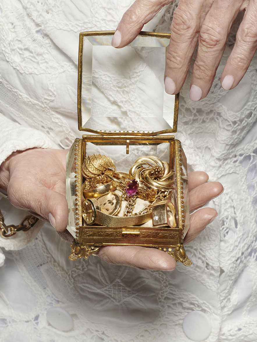 Nærbilde av en gammel dame i hvit kjole som holder et gjennomsiktig smykkeskrin med forskjellige gullsmykker i